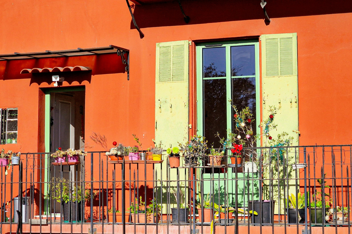 Balcone con numerosi vasi di fiori e piante su facciata arancione con persiane chiare.